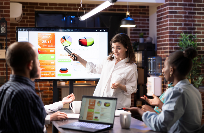 A woman giving a business analytics presentation to a diverse team in a modern office, using a large screen displaying charts and graphs.